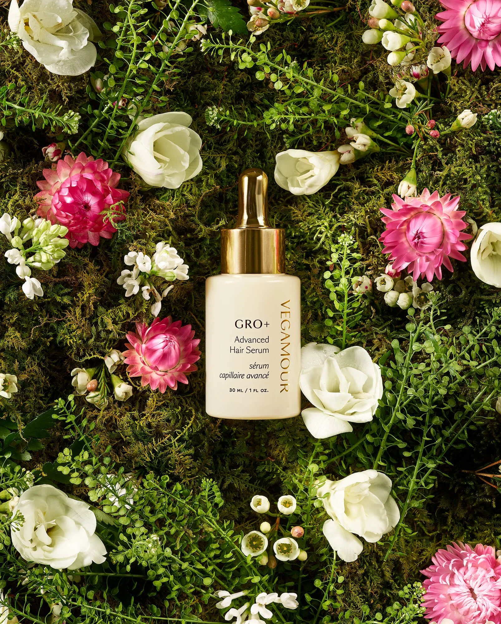 Bottle of GRO+ Advanced Hair Serum by Vegamour placed on green moss and surrounded by white and pink flowers.