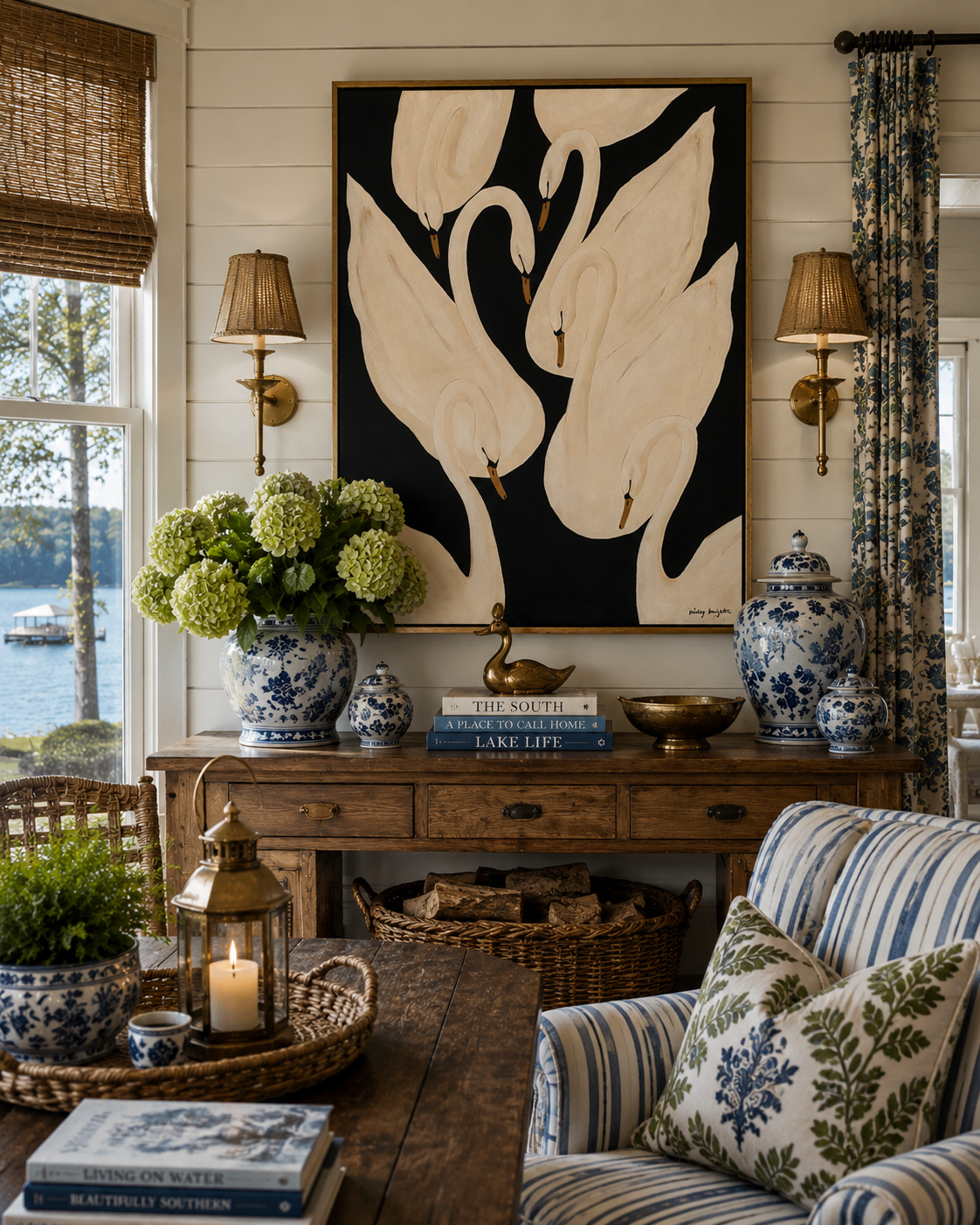 A cozy interior corner featuring a wooden sideboard decorated with blue and white porcelain vases, books, and a small brass duck figurine. Above the sideboard hangs a large painting of swans on a black background. Flanking the painting are wall-mounted lamps with woven shades. There is a vase with green hydrangeas, a basket of firewood, and a striped armchair with floral pillows in the foreground, with a window showing a lake and trees outside.