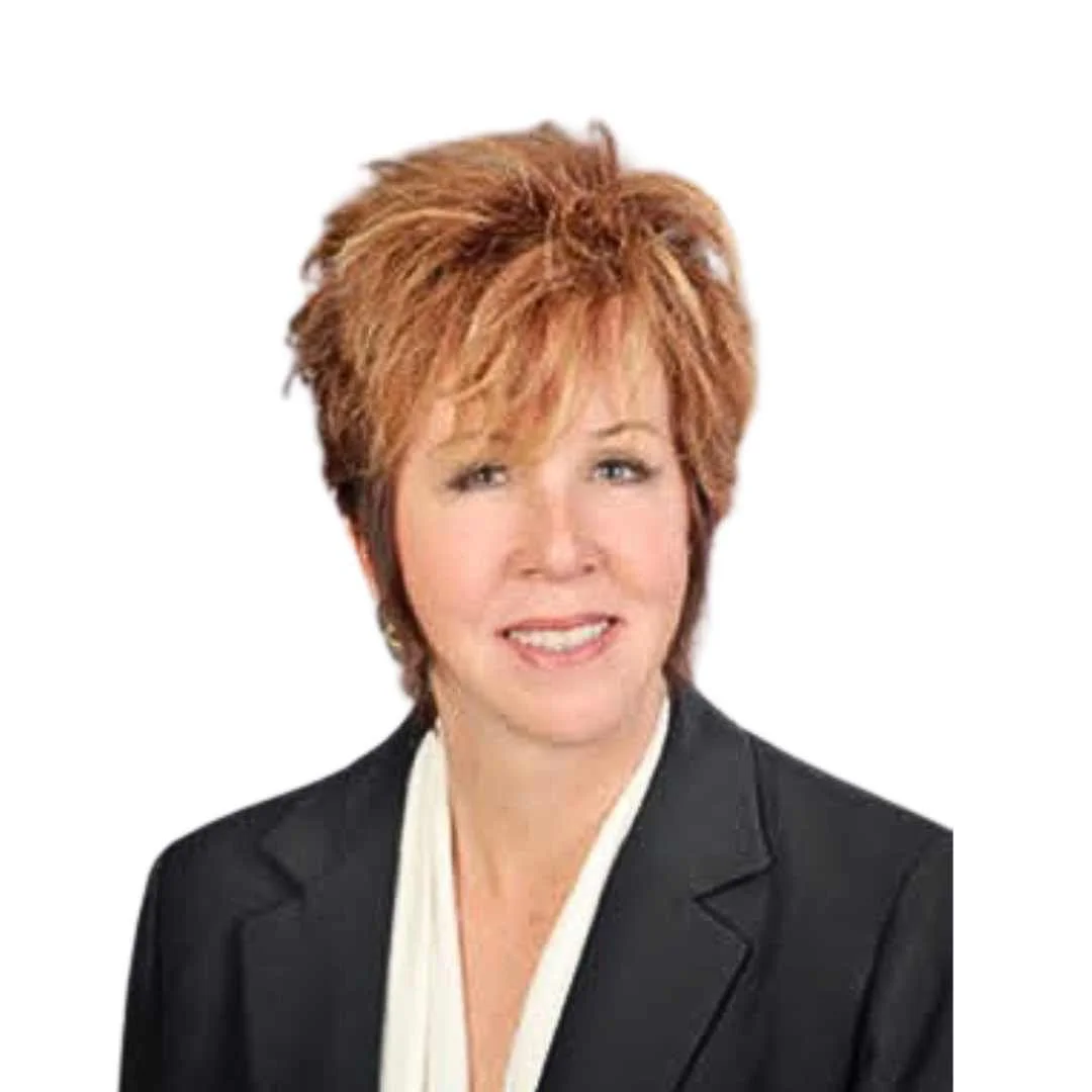 Portrait of a woman with short, reddish-brown hair, wearing a black blazer and a white top, smiling lightly against a plain white background.