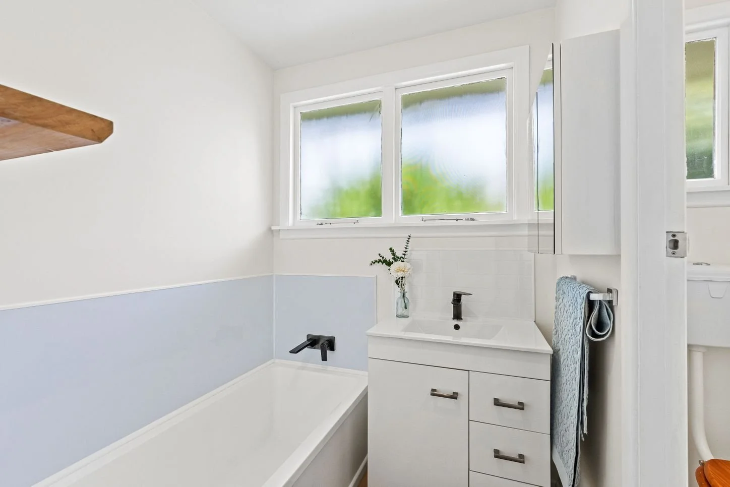 Bright bathroom with white walls, a bathtub, a small white vanity with a black faucet, a towel rack with towels, and a window with frosted glass.