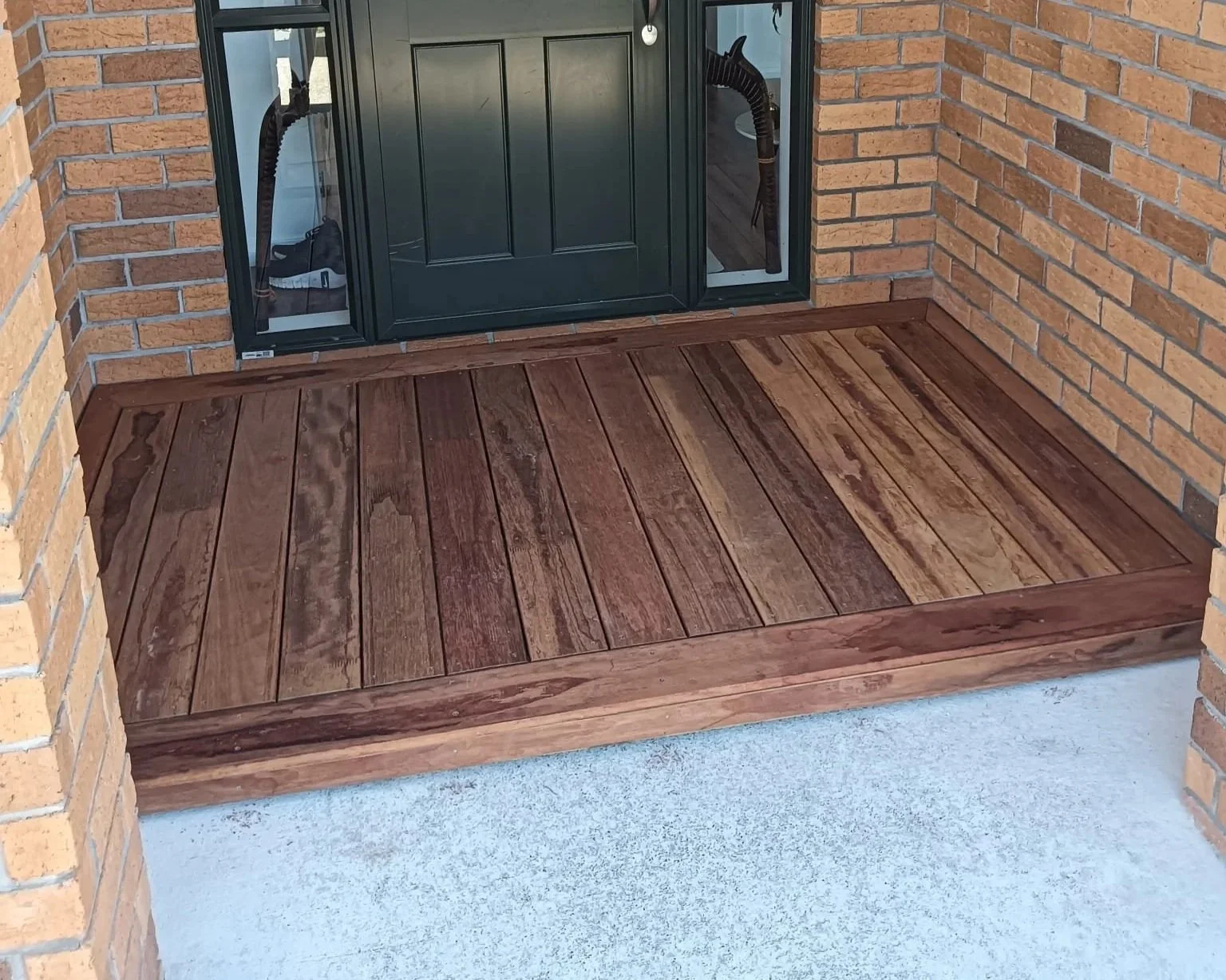 A small wooden deck with reddish-brown planks surrounded by brick walls, positioned in front of a black front door with glass sides.