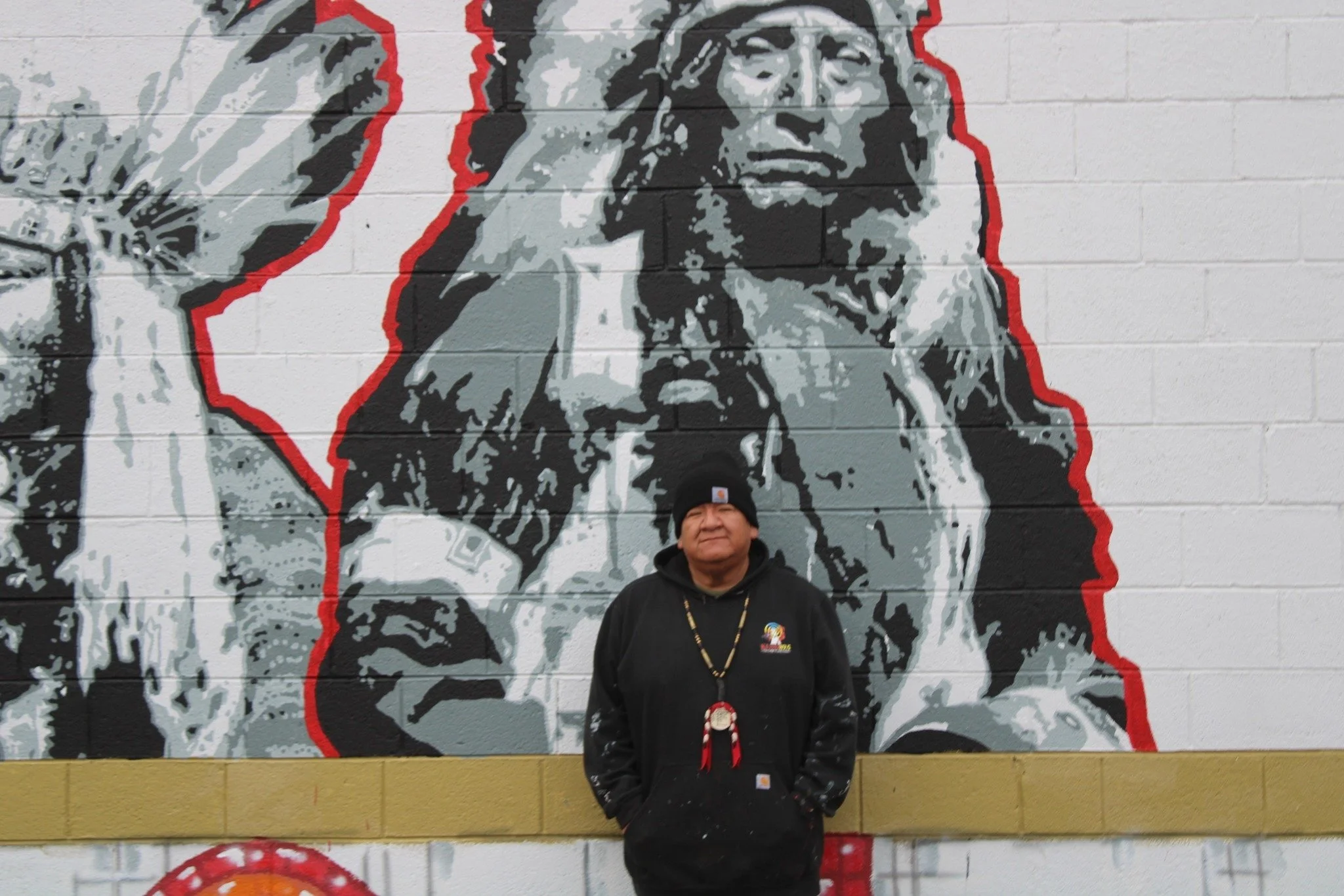 Person wearing a black Carhartt hoodie and a black beanie standing in front of a mural of a Native American chief on a brick wall. The person is smiling with hands in pockets, and is wearing a beaded necklace and a red ribbon medal.