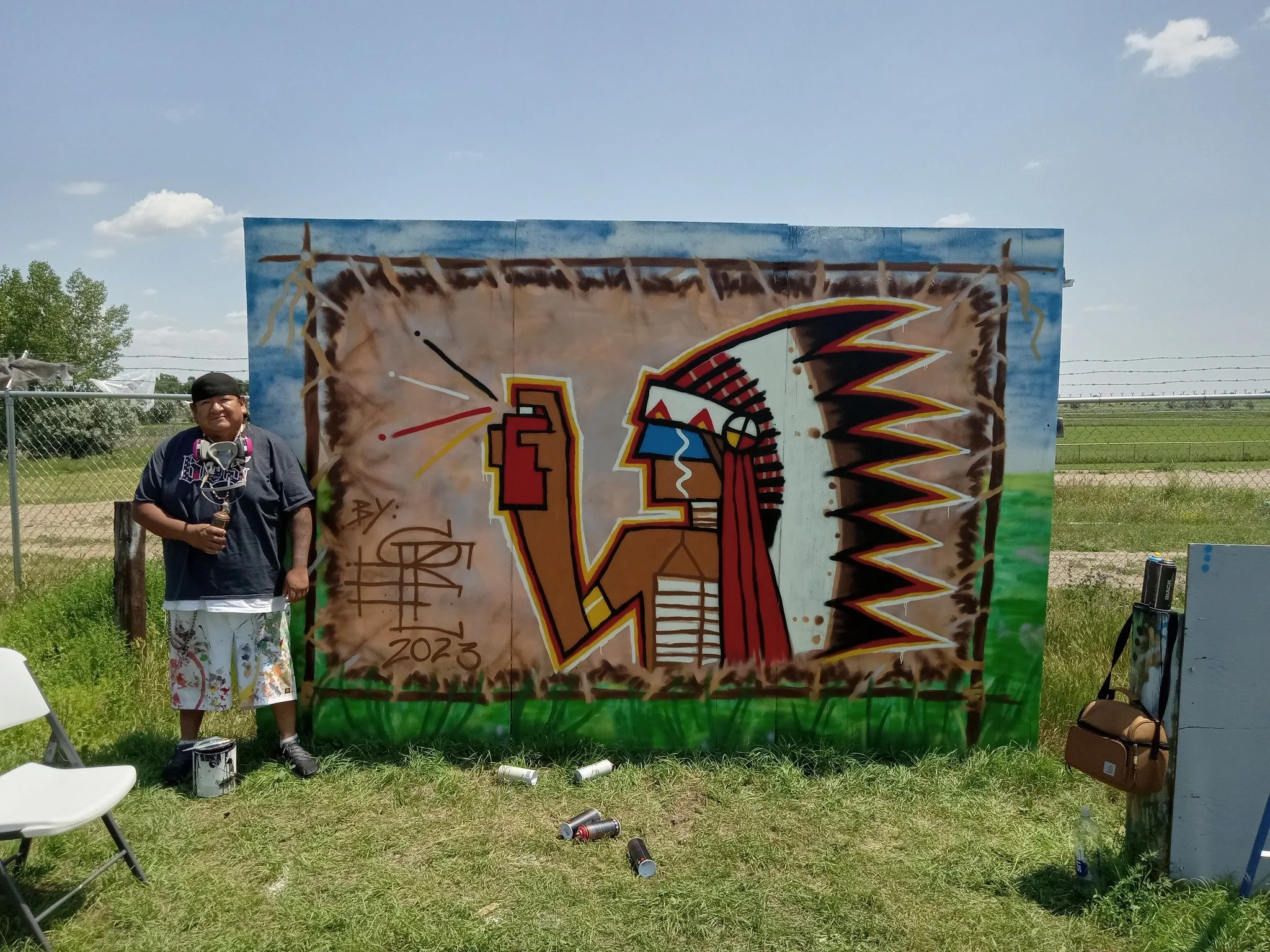 A man standing next to a colorful mural of a Native American chief with a feathered headdress and a landscape background, outdoors on a sunny day.