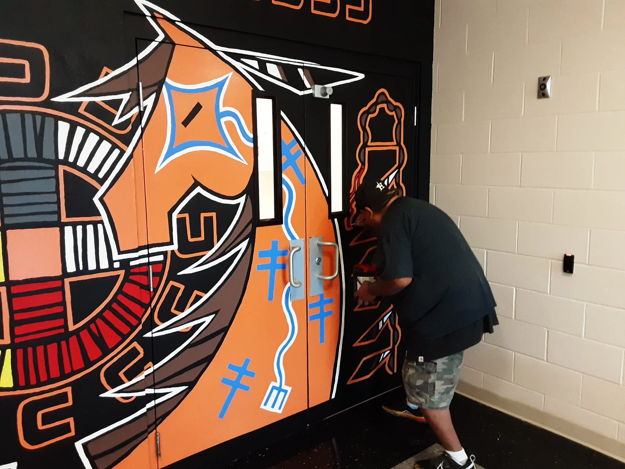 A person in black shirt and camouflage shorts installing or fixing a colorful mural on a door, featuring orange, blue, white, and black abstract shapes and symbols.