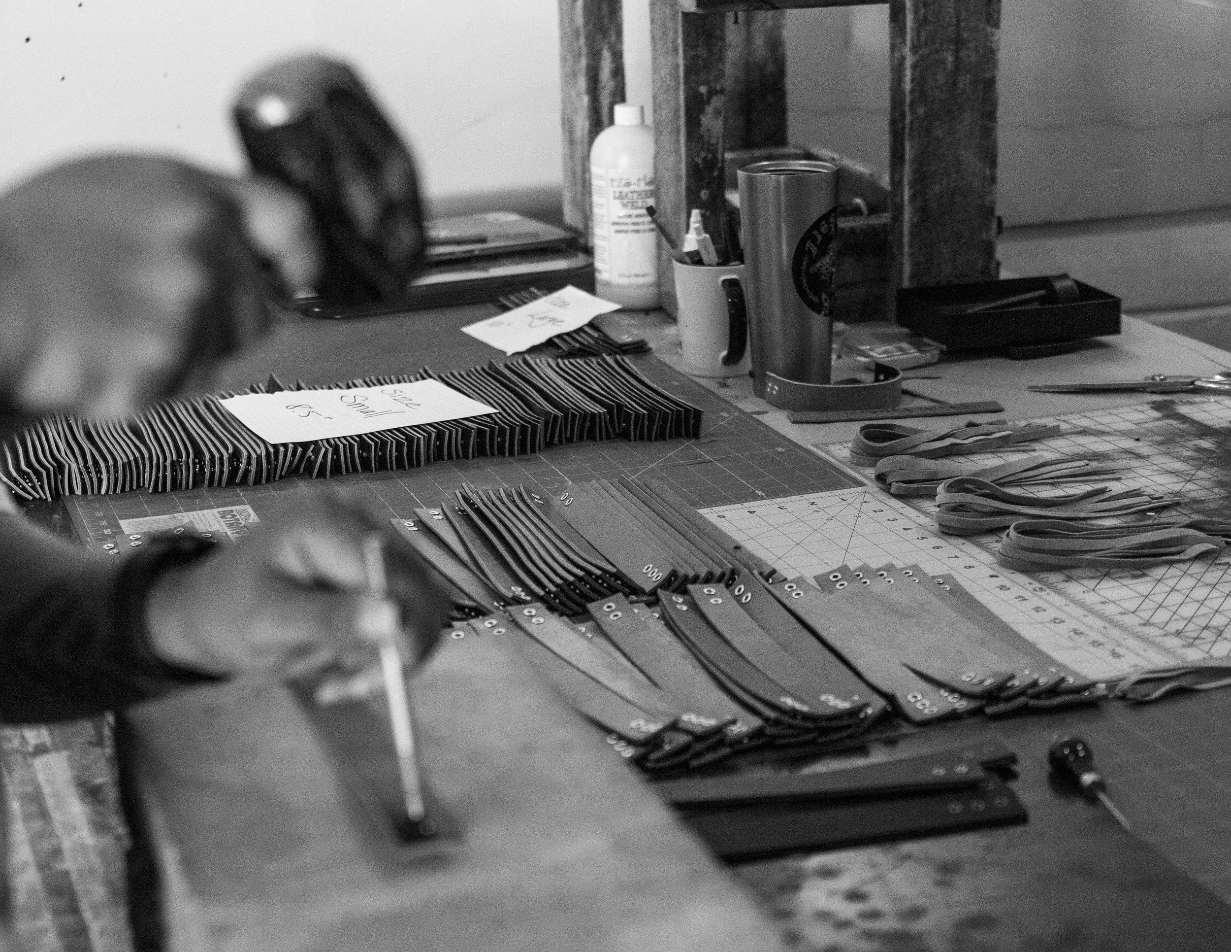 Person working on leather crafting, using a tool on leather strips with grommets on a work table, accompanied by various crafting tools, a cutting mat, and a tumbler.