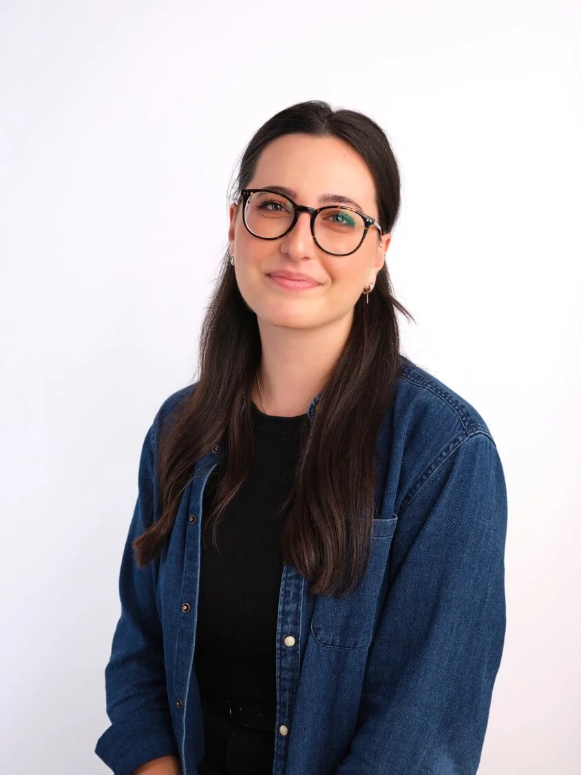 Headshot of a young woman with long brown hair wearing glasses, a black shirt, and a denim jacket, smiling and looking at the camera against a white background.