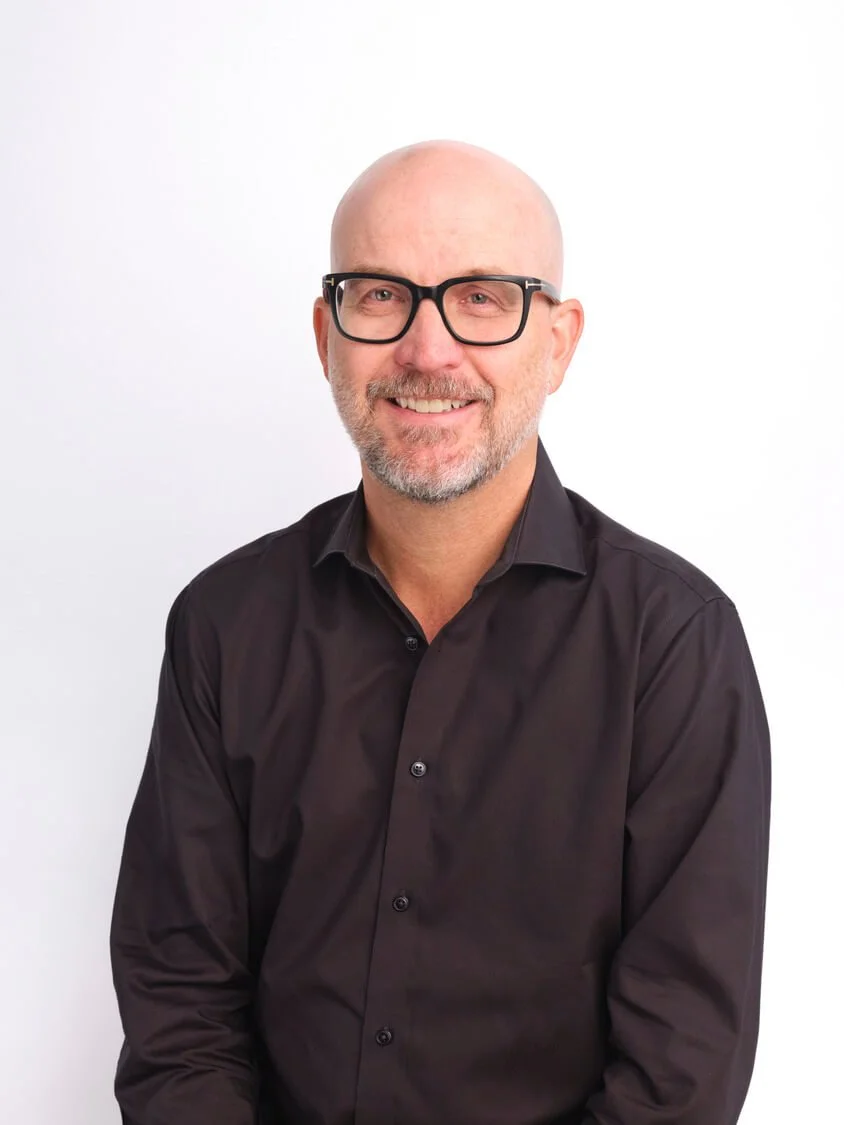 A bald man with glasses and a beard smiling, wearing a black shirt, standing against a white background.
