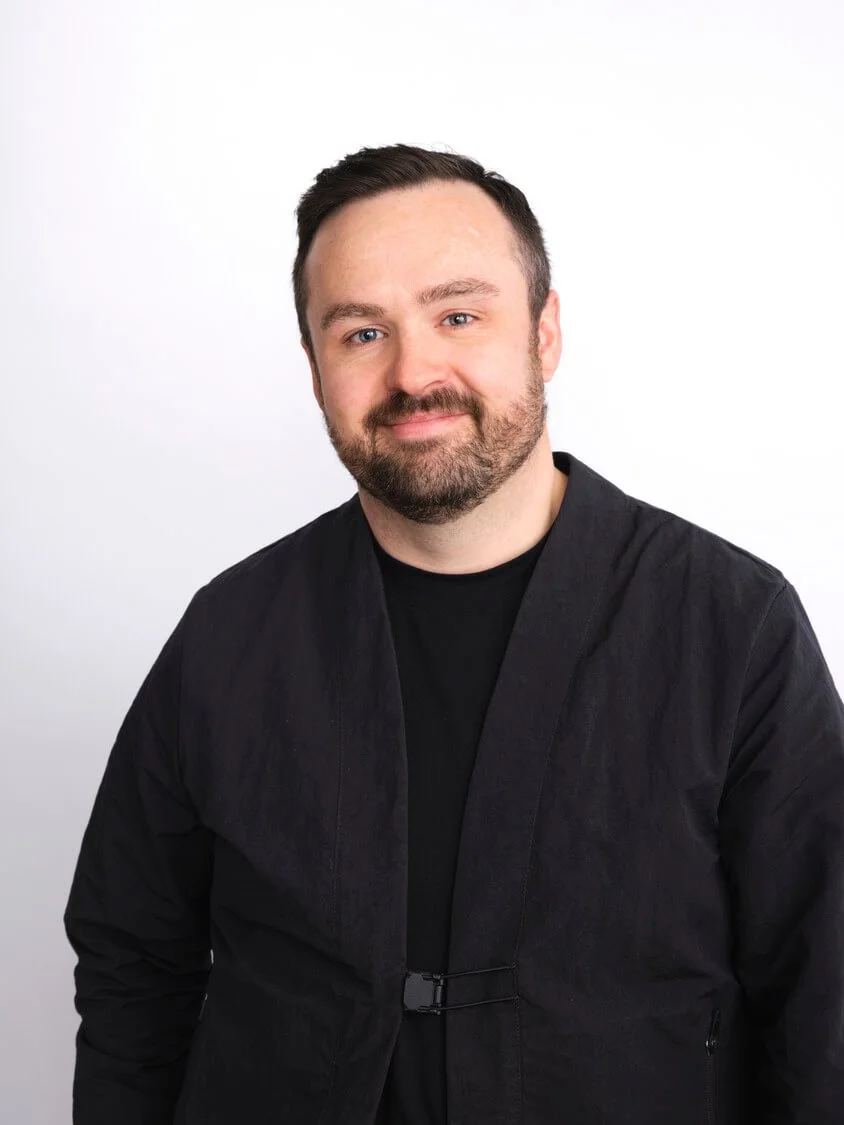 Headshot of a man with short dark hair and a beard, smiling, wearing a black jacket over a black shirt, standing against a plain white background.