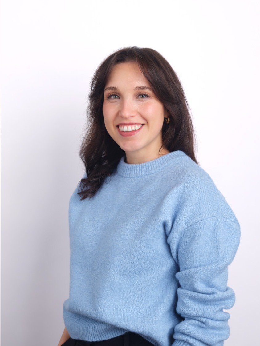 Headshot of a smiling woman with brown hair wearing a light blue sweater against a white background.