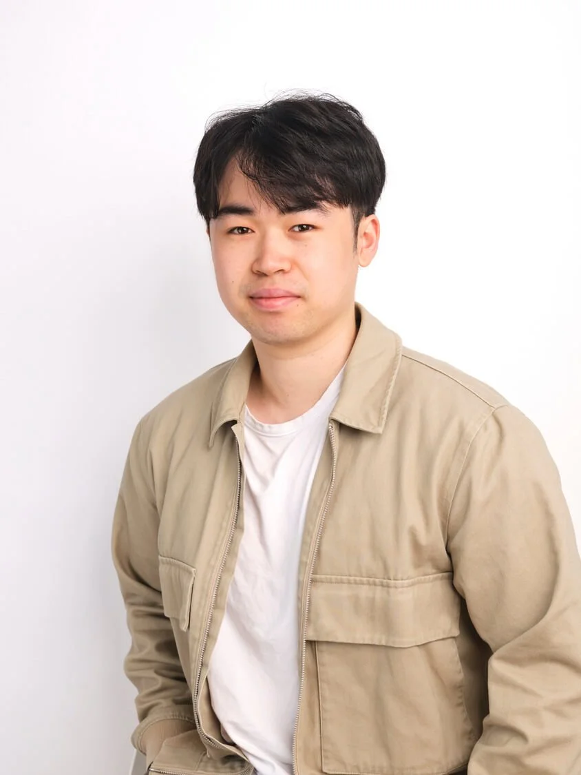 Headshot of a young man with black hair, wearing a beige jacket over a white t-shirt, standing against a plain white background.