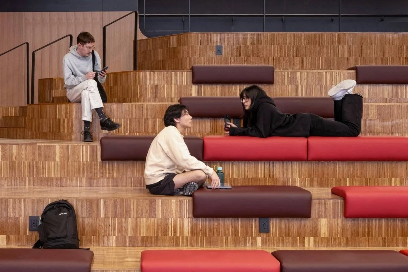 Three students with backpacks engaging with their phones on tiered wooden seating in a modern indoor space, representing campus projects