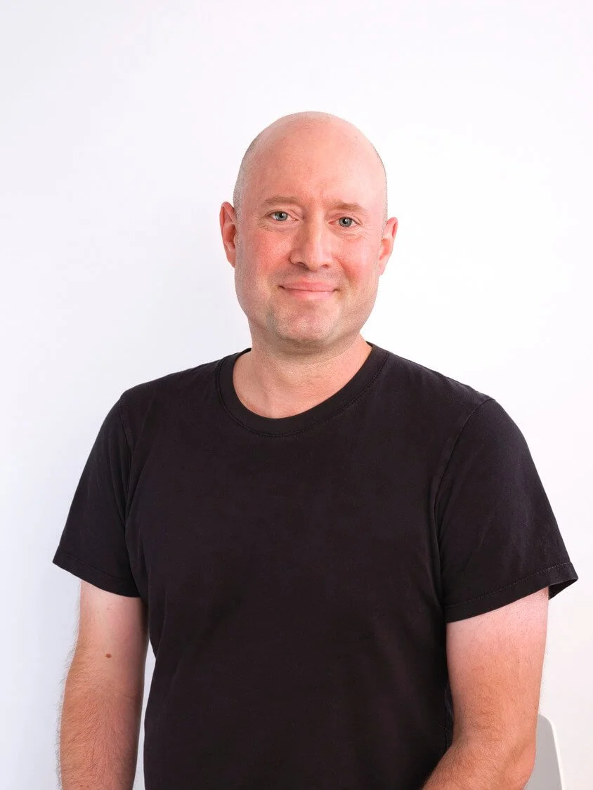 Headshot of a man with a shaved head, wearing a black T-shirt, standing against a plain white background, smiling softly at the camera.