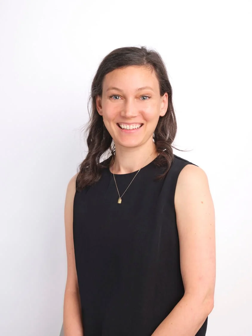 Headshot of a woman with shoulder-length brown hair, wearing a black sleeveless top and a gold necklace, smiling at the camera against a plain white background.