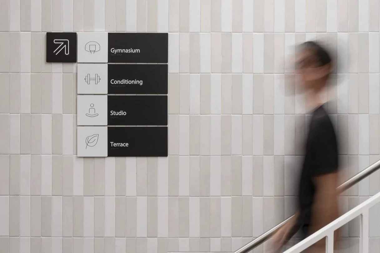 A blurry person walking past a black and white directional sign on a tiled wall, indicating locations like Gymnasium, Conditioning, Studio, and Terrace.