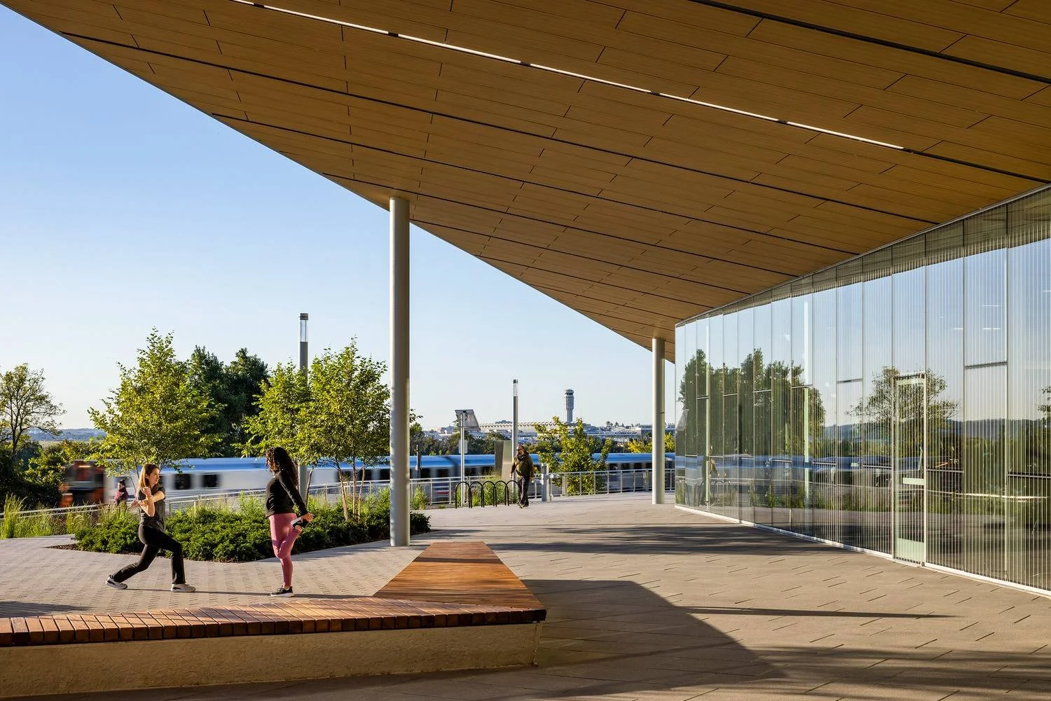 People walking outside a modern building with a large overhanging roof, glass walls, trees, and a train passing by in the background.