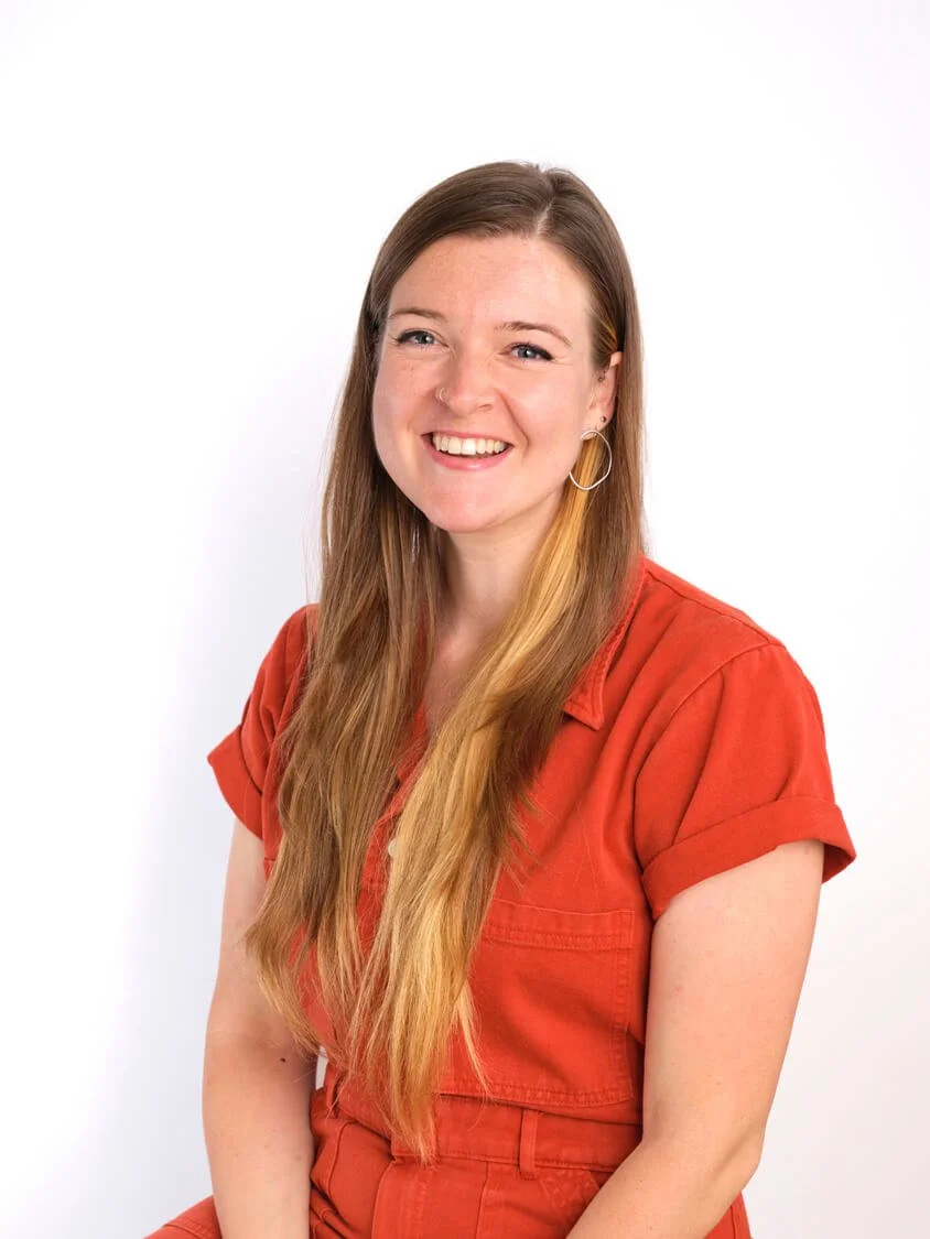Headshot of a woman with long, light brown hair wearing an orange shirt, smiling, and wearing hoop earrings against a white background.