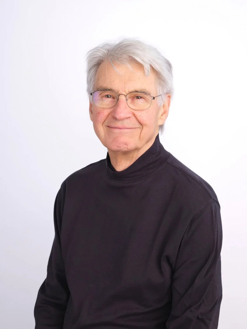 Headshot of an older man with grey hair, glasses, wearing a black turtleneck, smiling against a plain white background.