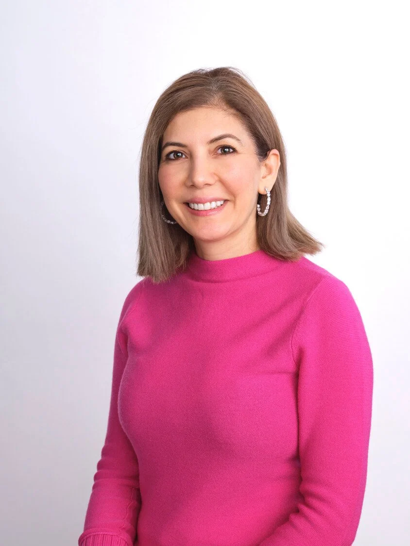 Headshot of a woman with shoulder-length brown hair wearing a pink long-sleeve top and hoop earrings, smiling at the camera against a white background.