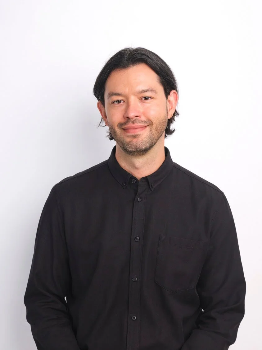 Headshot of a man with dark hair and a slight beard, wearing a black button-down shirt, standing against a white background with a slight smile.