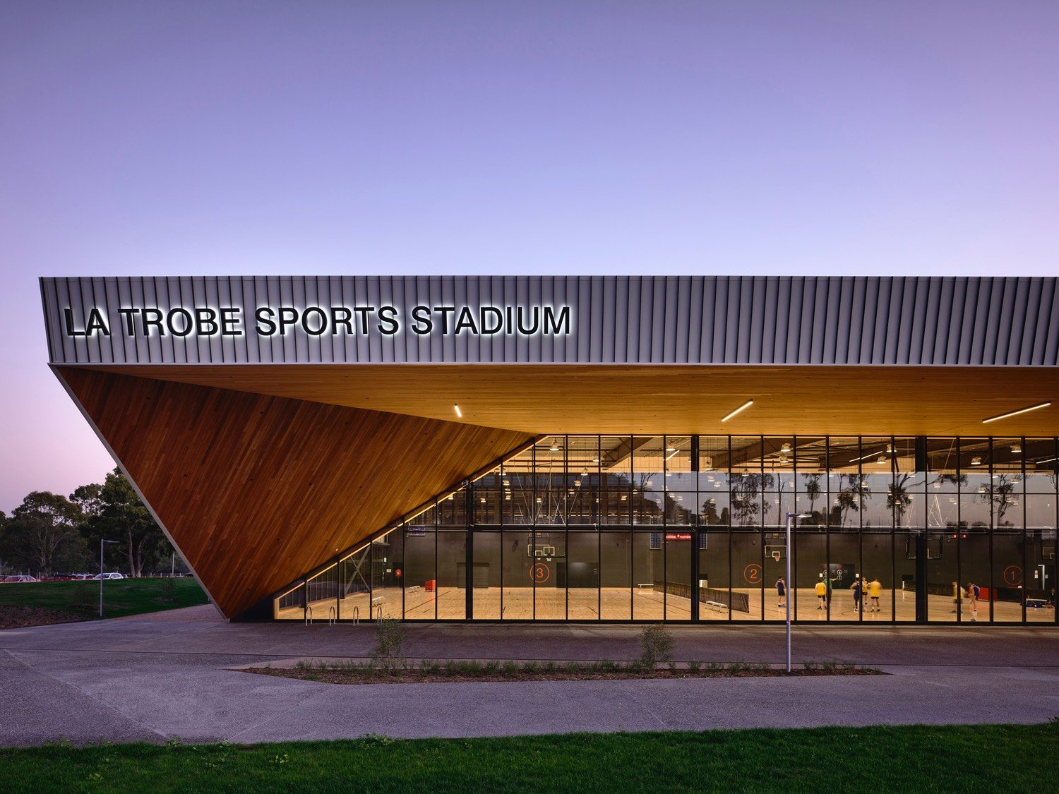 Exterior of La Trobe Sports Stadium with large glass windows showing a basketball court inside, modern wooden and metal architectural design, during dusk with a purple sky.