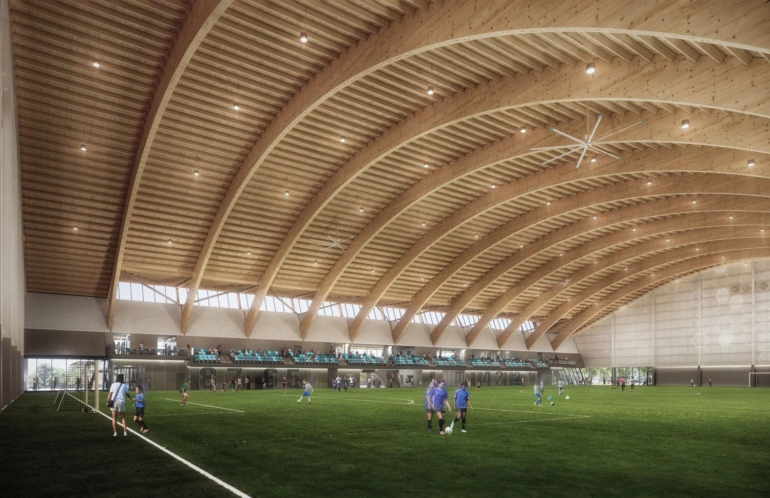 Indoor soccer facility with a wooden arched ceiling, green artificial turf, children playing soccer, and spectators seated in the stands.