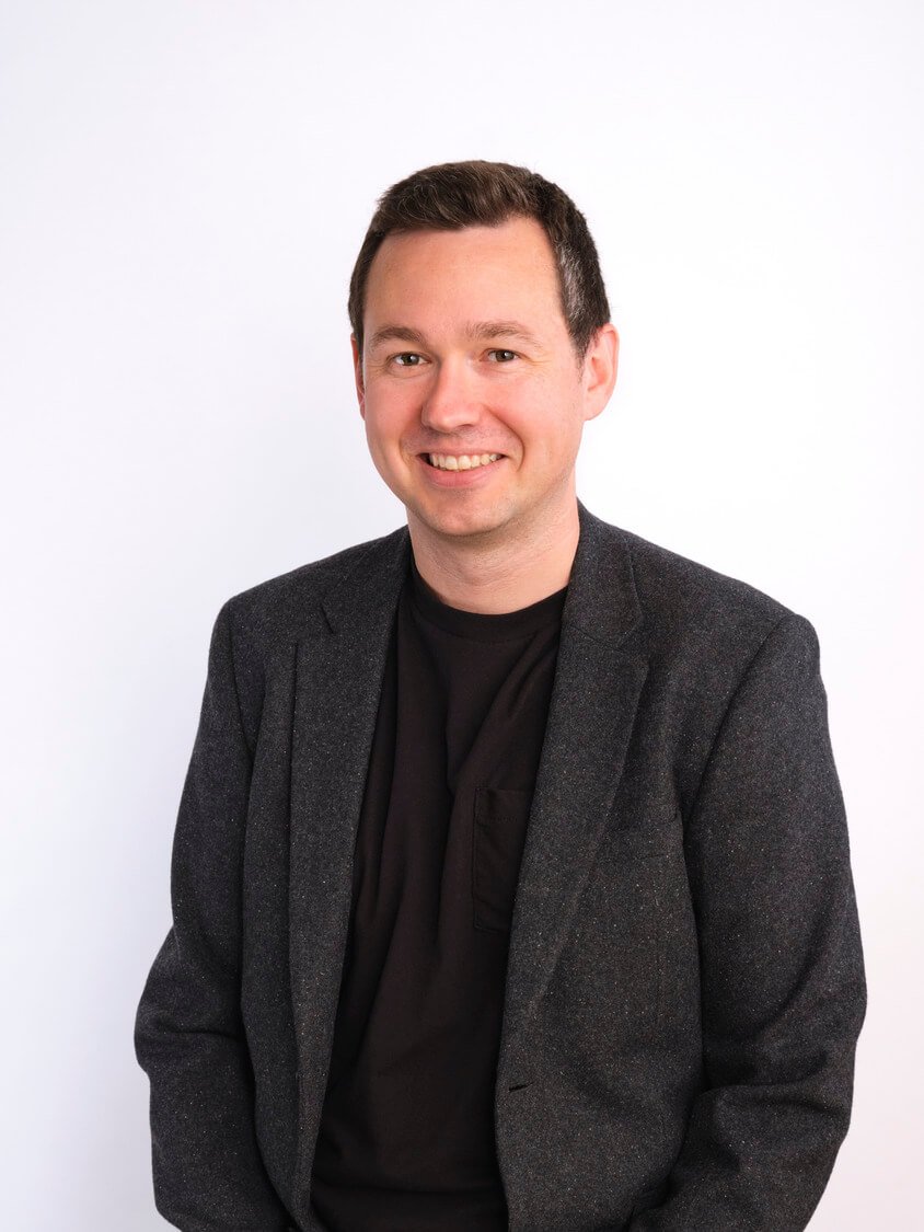 Headshot of a smiling man with short dark hair, wearing a black shirt and dark grey blazer, standing against a plain white background.