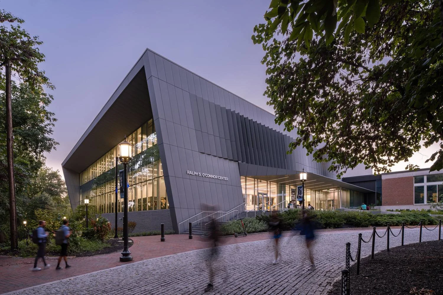 Exterior view of the Ralphs O'Connor Center with modern architectural design, large glass windows, and a slanted roof, during dusk with people walking and trees visible.