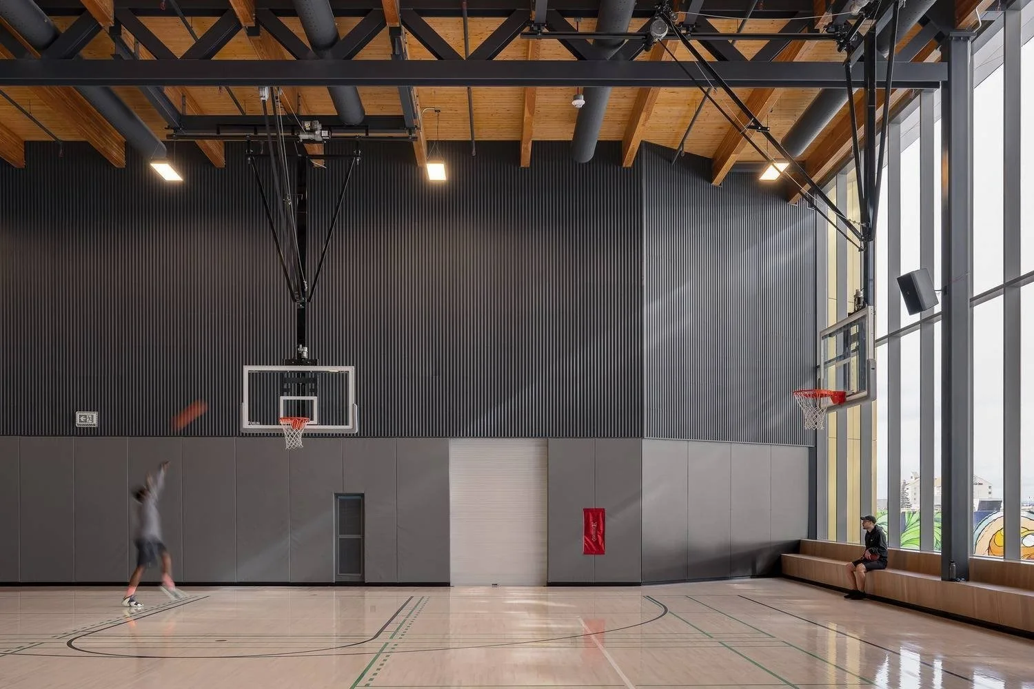 Indoor basketball court with two hoops, one of which is in use by a person jumping to shoot a basketball, and another person sitting on a wooden bench near large windows.
