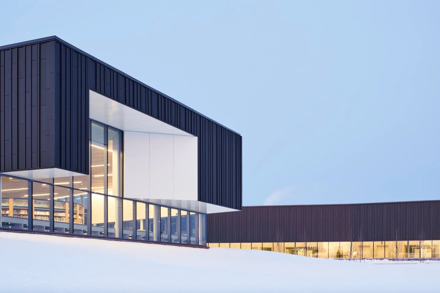 Modern library building with black exterior, large glass windows, and interior lighting, surrounded by snow under a clear blue sky.
