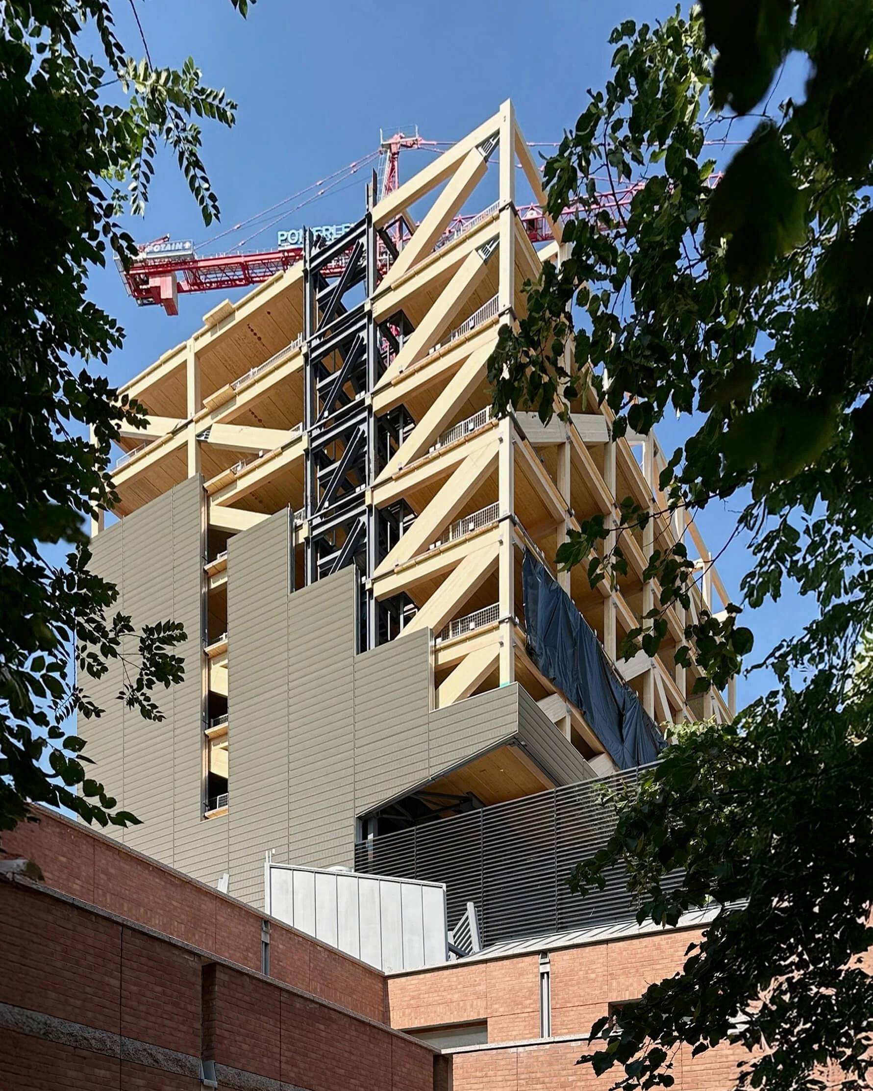 A construction site of a modern building with wooden and metal framework, surrounded by trees and blue sky.