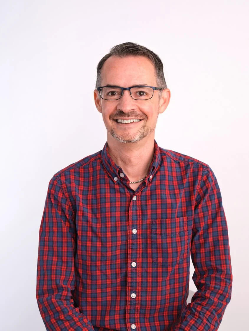 Headshot of a smiling man with glasses and a goatee, wearing a red and blue plaid shirt, standing against a plain white background.