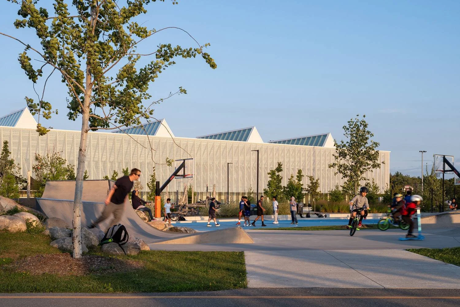 People skateboarding and riding bikes at a skate park with a modern building in the background on a clear day.
