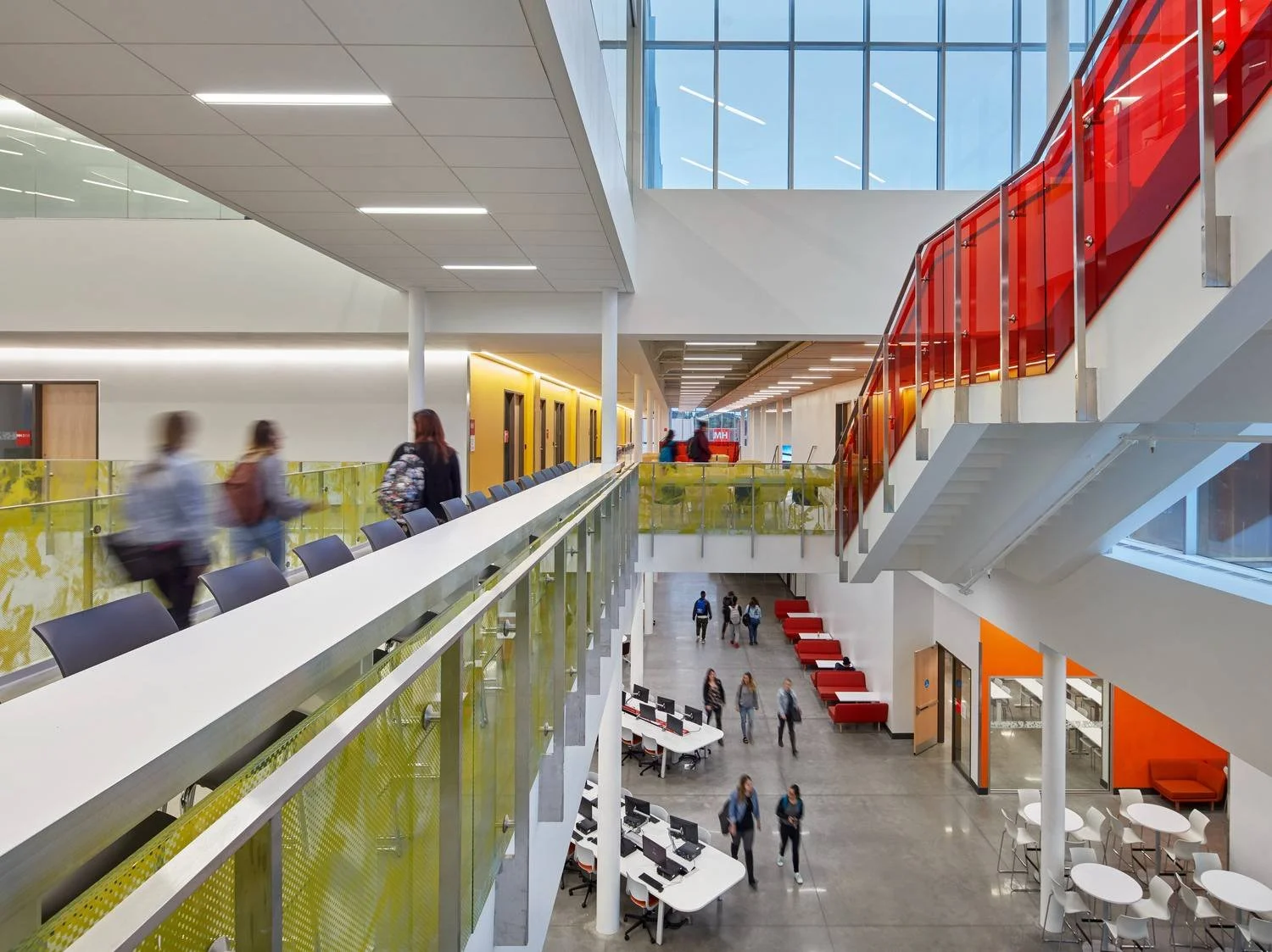 Interior of a modern, multi-level educational or office building with people walking and sitting. The image features a glass-paneled upper walkway with yellow and red accents and a staircase with a red glass railing.
