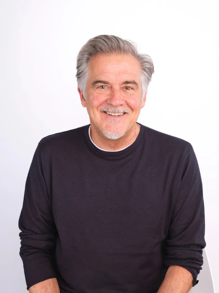 Headshot of a smiling older man with grey hair and beard, wearing a black sweater, sitting against a plain white background.