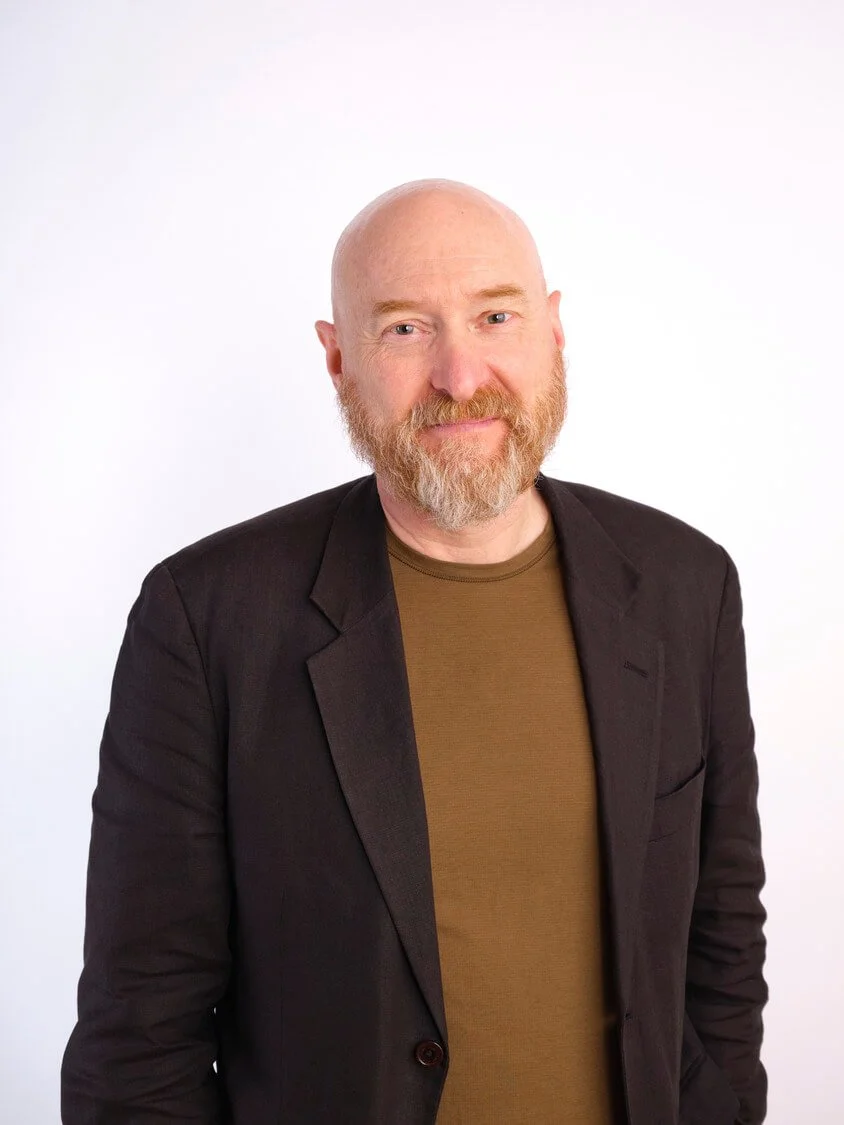 A bald man with a beard wearing a brown shirt and black blazer posing against a white background.