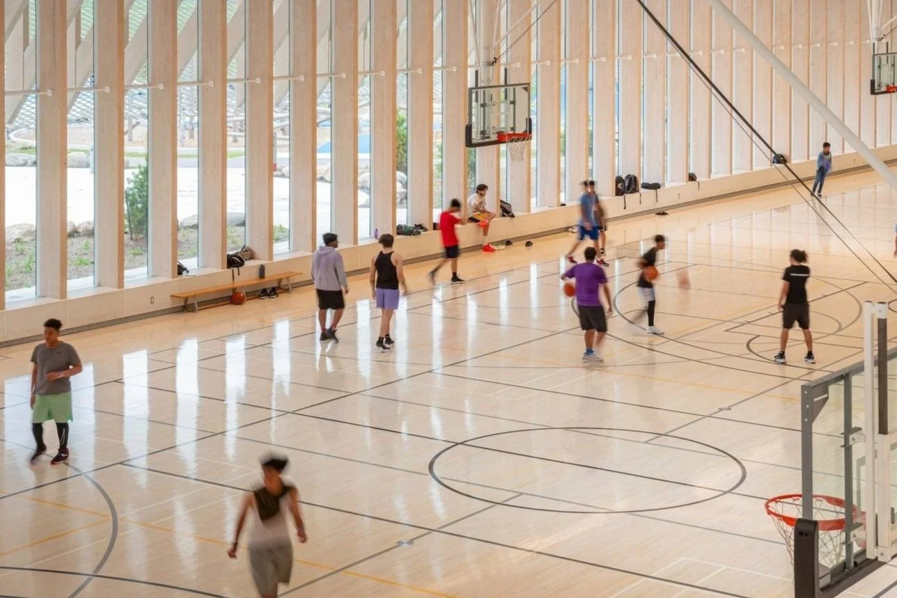 People playing basketball in an indoor gym with large windows, hardwood floor, and basketball hoops to represent community projects