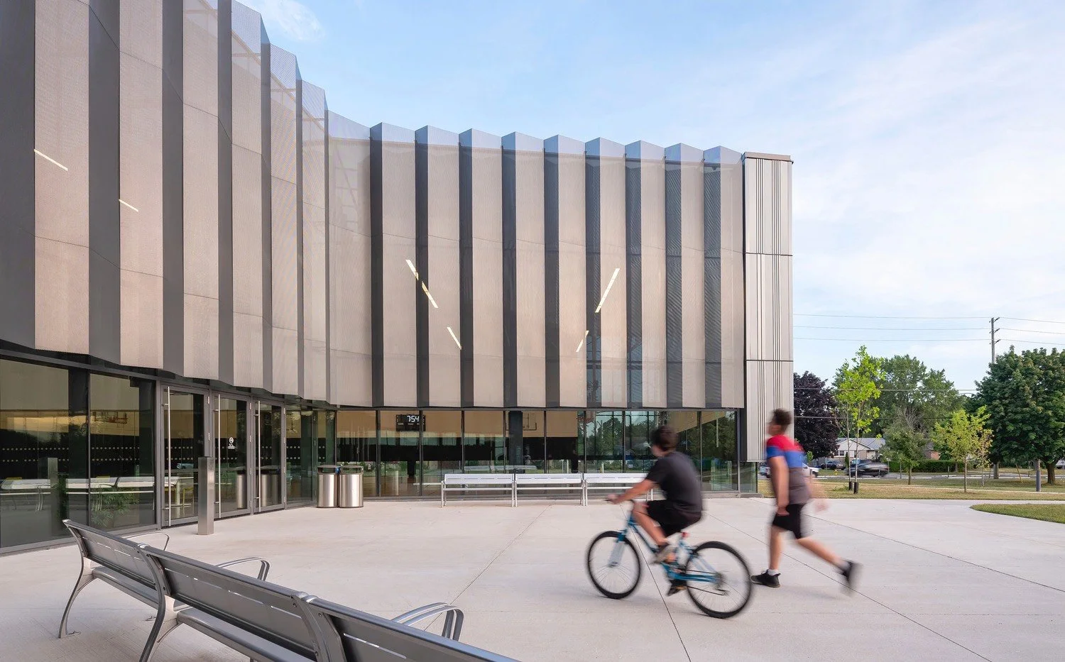 Modern building with metallic facade and large glass entrance, outdoor plaza with benches, a person riding a bicycle, and another person walking, trees and blue sky in the background.
