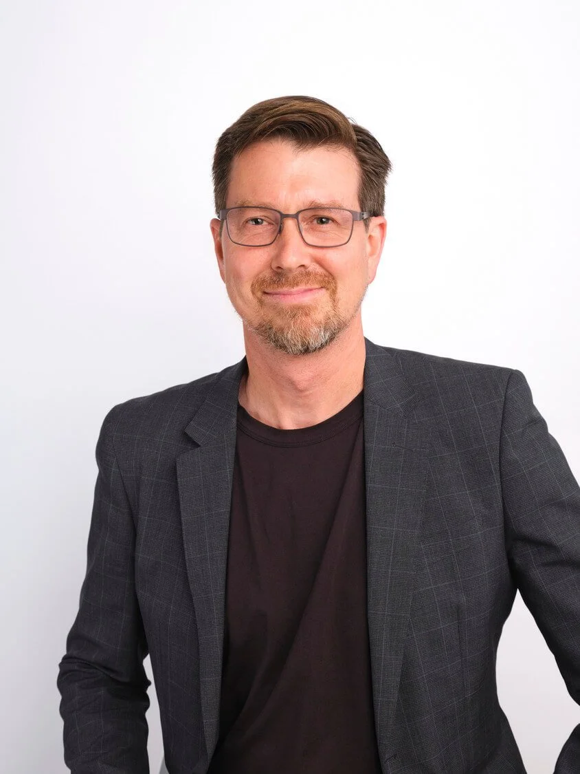 A man with glasses, a beard, and brown hair wearing a black shirt and a dark blazer, smiling at the camera against a white background.
