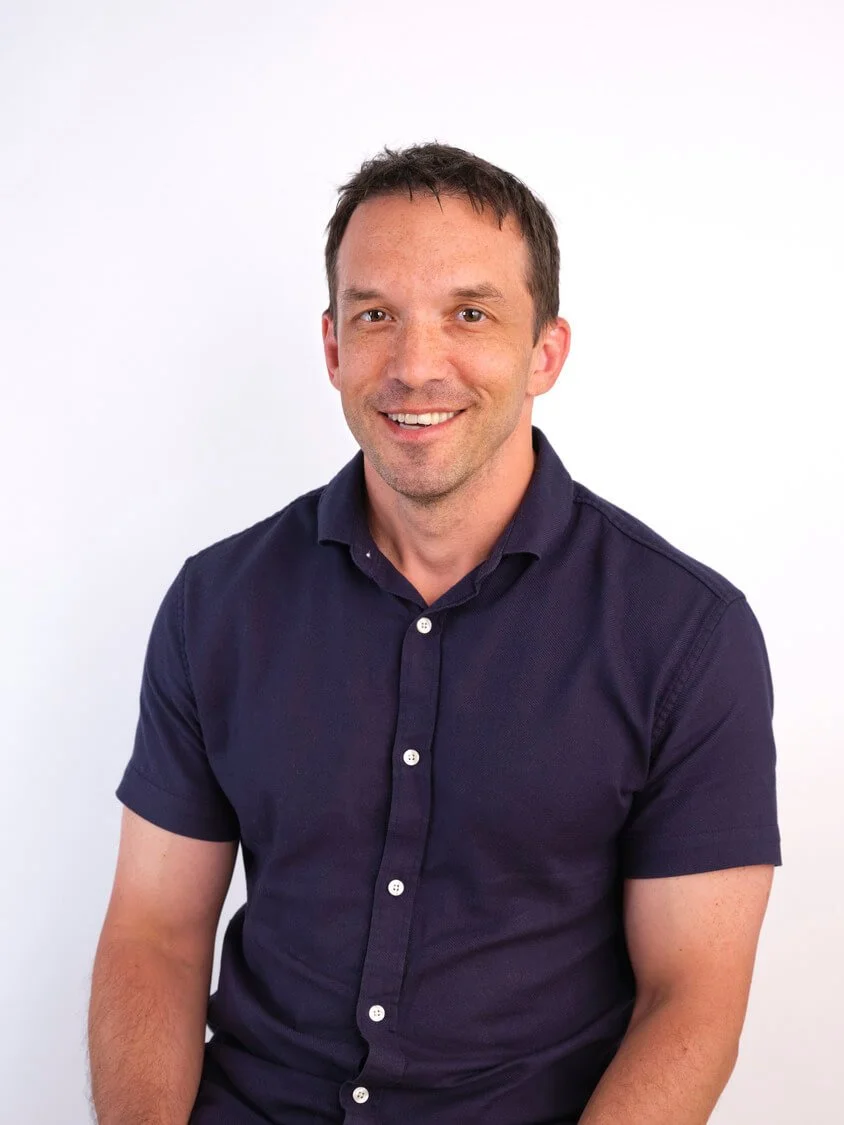 A smiling man with short dark hair wearing a navy blue collared shirt against a plain white background.
