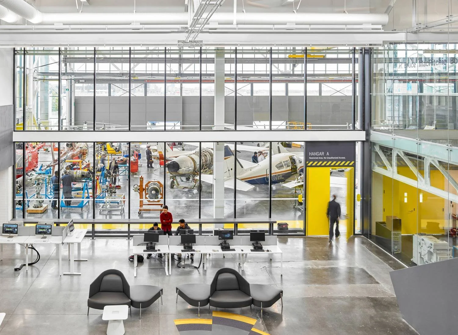 View of a lobby with seating areas and a few people using computers. An hangar with planes, equipment, and students is seen through large glass windows.