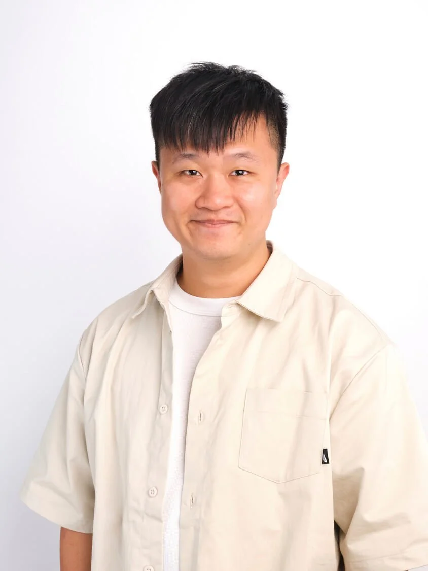 Headshot of a man with black hair smiling, wearing a beige button-up shirt over a white t-shirt, against a plain white background.