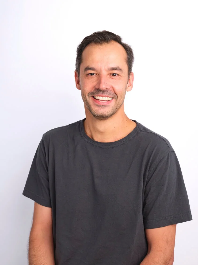 Headshot of a smiling man with short dark hair, wearing a black t-shirt, standing against a plain white background.