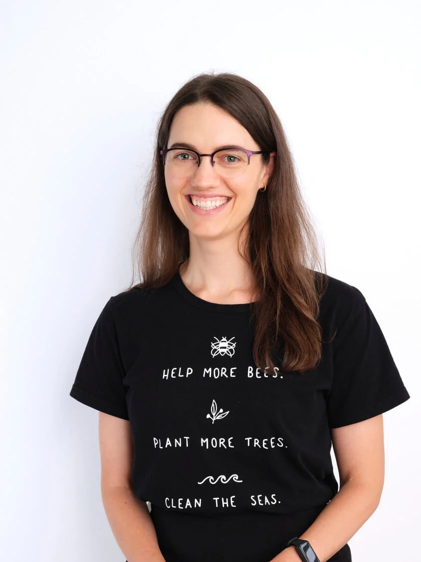 Headshot of a woman with long brown hair, wearing glasses and a black t-shirt that says 'Help More Bees. Plant More Trees. Clean the Seas.' She is smiling and standing against a plain white background.