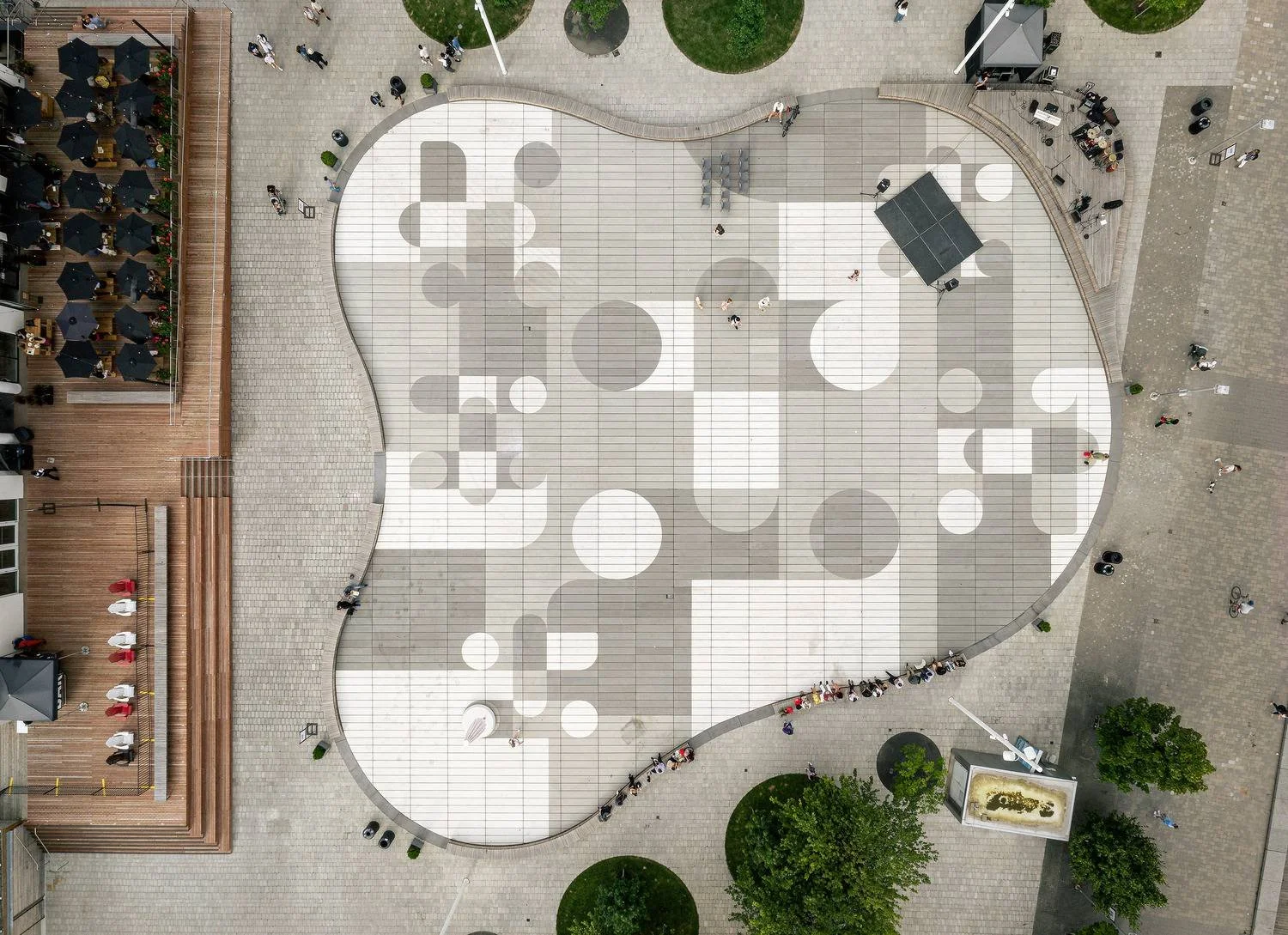An aerial view of an outdoor plaza with geometric patterns on the pavement, surrounded by people, trees, and a building with a rooftop patio.
