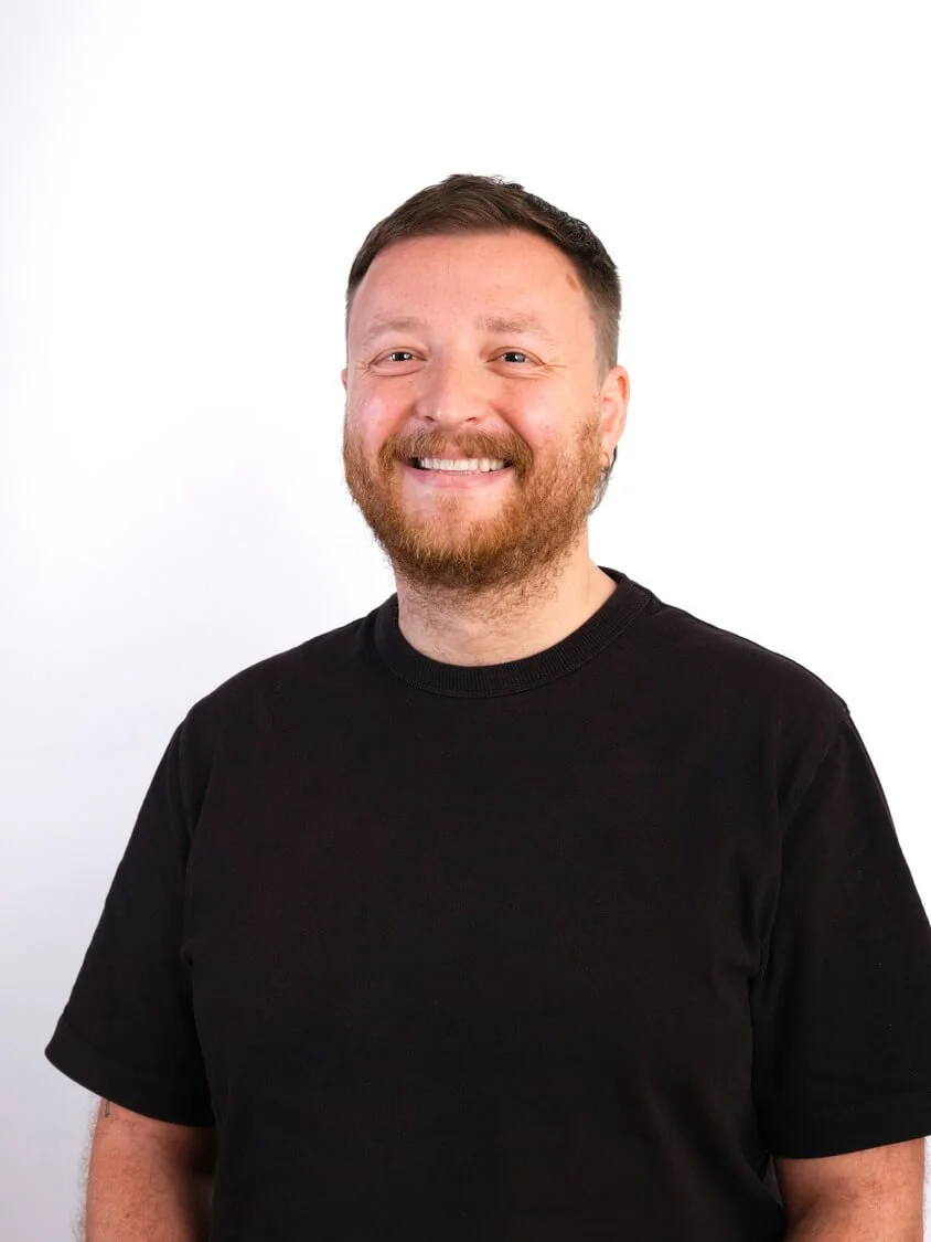Headshot of a smiling man with short brown hair, a beard, and wearing a black t-shirt, standing against a white background.