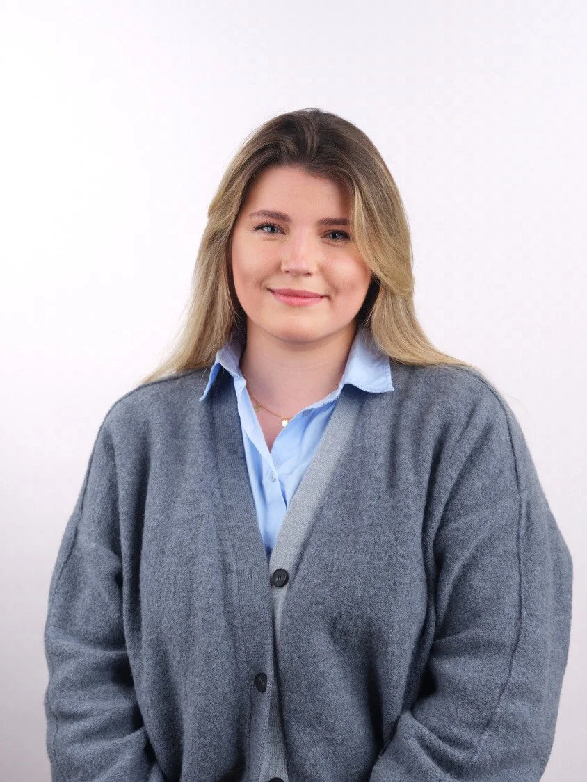 Headshot of a young woman with blonde hair wearing a blue collared shirt and a grey cardigan, standing against a plain white background, smiling slightly.