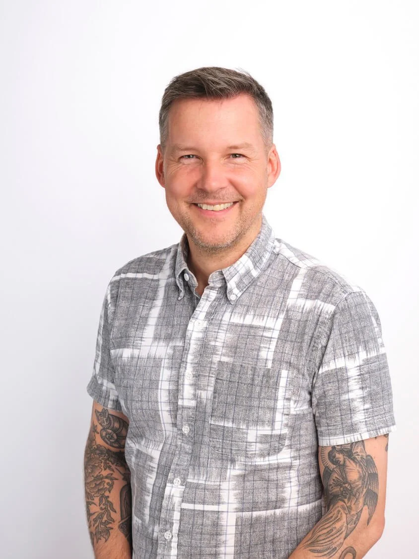 A smiling middle-aged man with short brown hair and tattoos on his arms, wearing a grey patterned button-up shirt, standing against a plain white background.