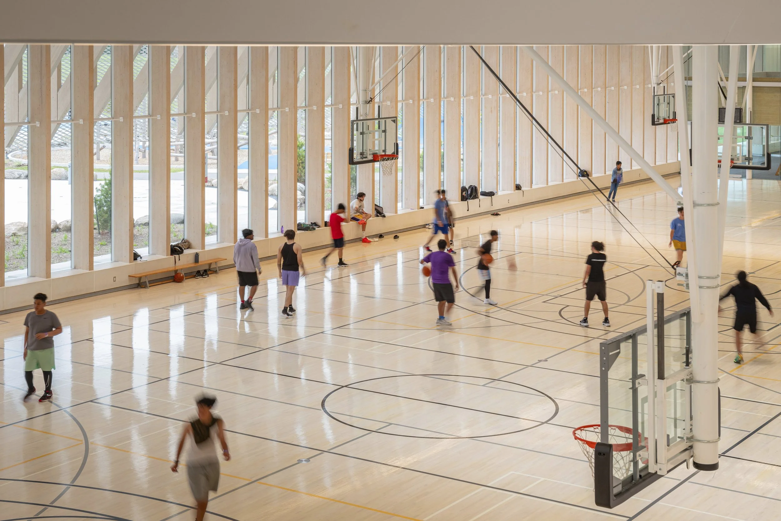 People playing basketball in an indoor gym with high wooden walls, multiple basketball hoops, and large windows.