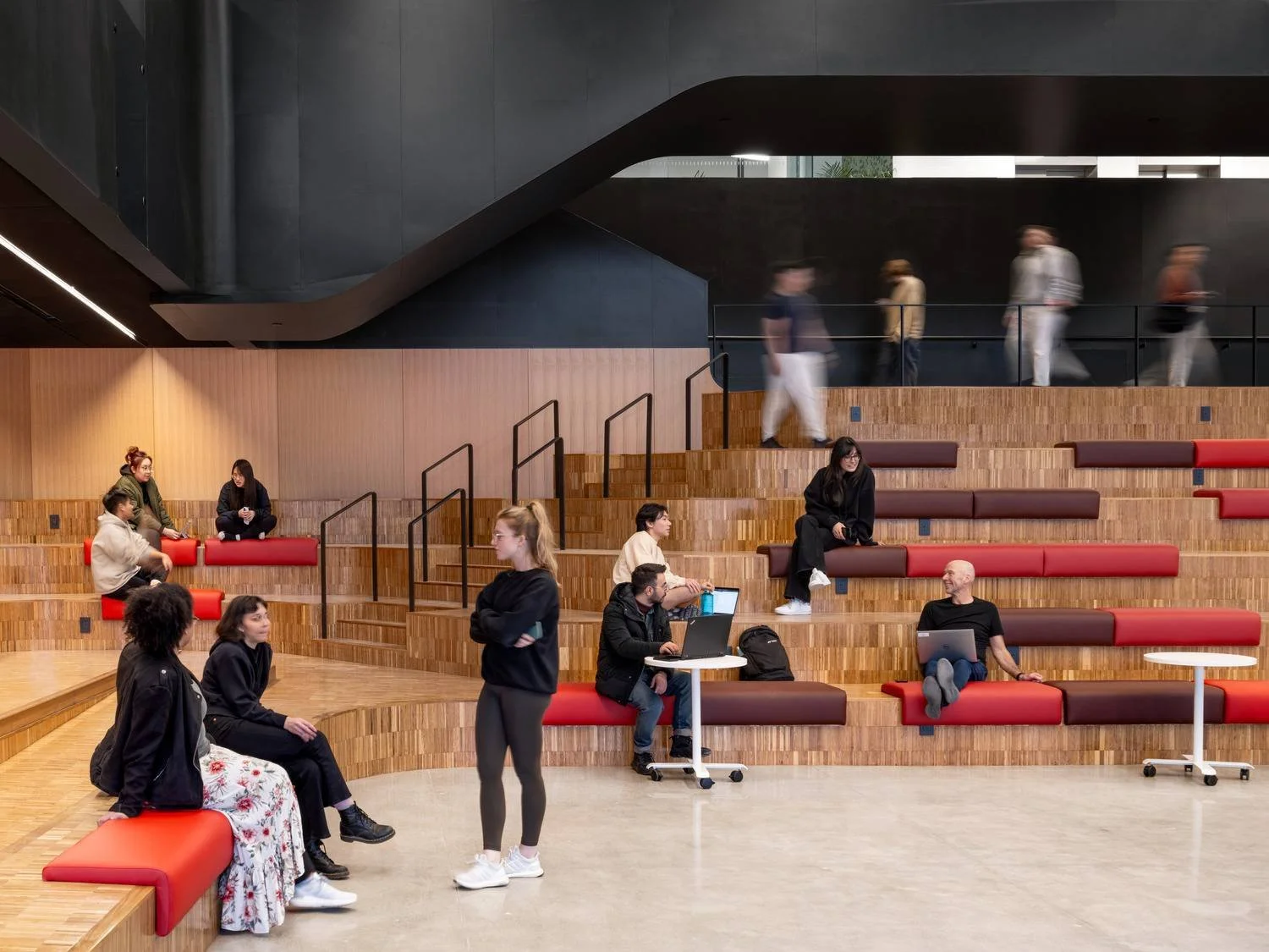 People seated on tiered wooden seating with red cushions in a modern, spacious interior, some working on laptops, others engaged in conversations, with a few walking along the upper level.