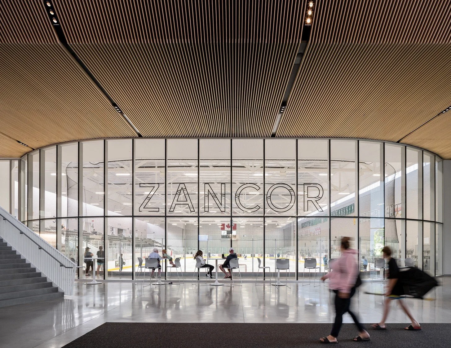 Interior of a modern ice skating rink with large glass front doors displaying the word ZANCOR. Several people are sitting and walking, with some active on the ice behind the glass. The ceiling has a wooden slat design.